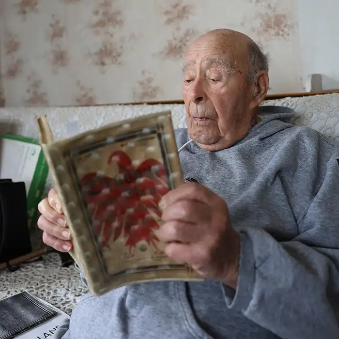 Cette photo est un portrait de Raymond en sweat gris tenant dans ses mains un carnet d'archives.